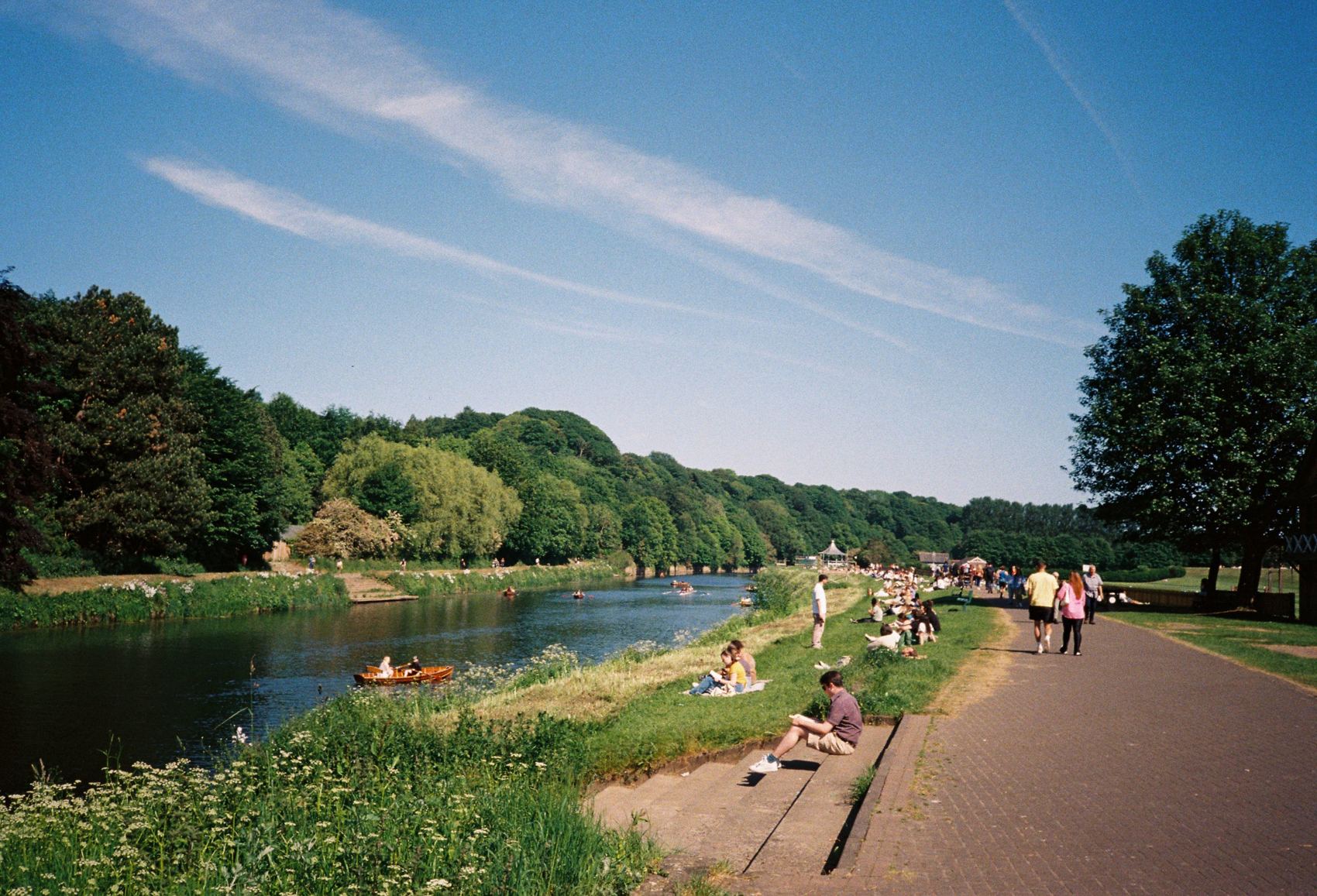 People reading on a riverbank under a streaked summer sky, with rowing boats on the water